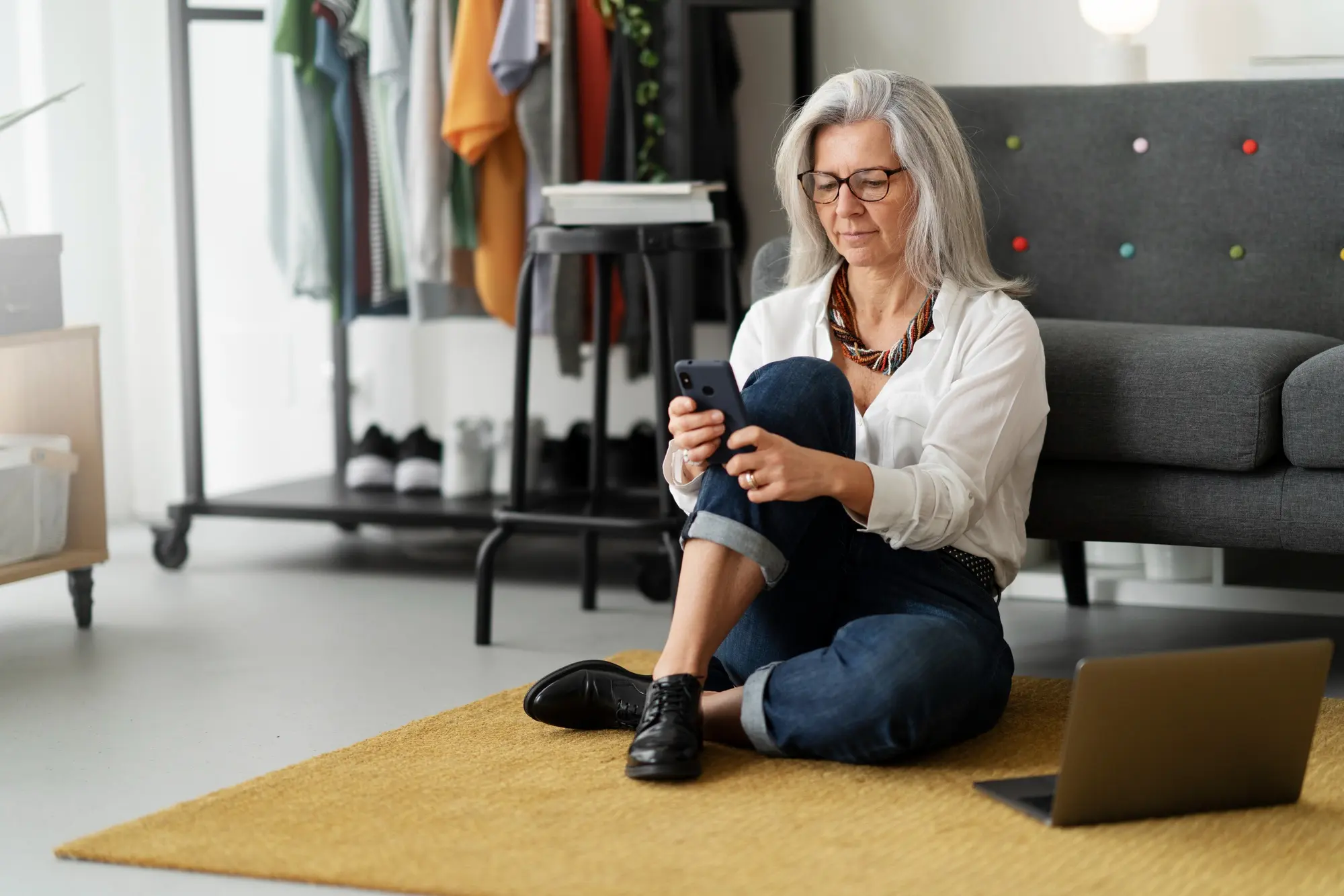 Frau Anfang 60 mit langem grauem Haar und Brille sitzt entspannt auf einem senfgelben Teppich am Boden, lehnt an einem grauen Sofa und schaut auf ihr Smartphone. Weiße Bluse, dunkle Jeans, ein Laptop neben ihr. Modernes, helles Wohnzimmer mit Kleiderstange im Hintergrund.