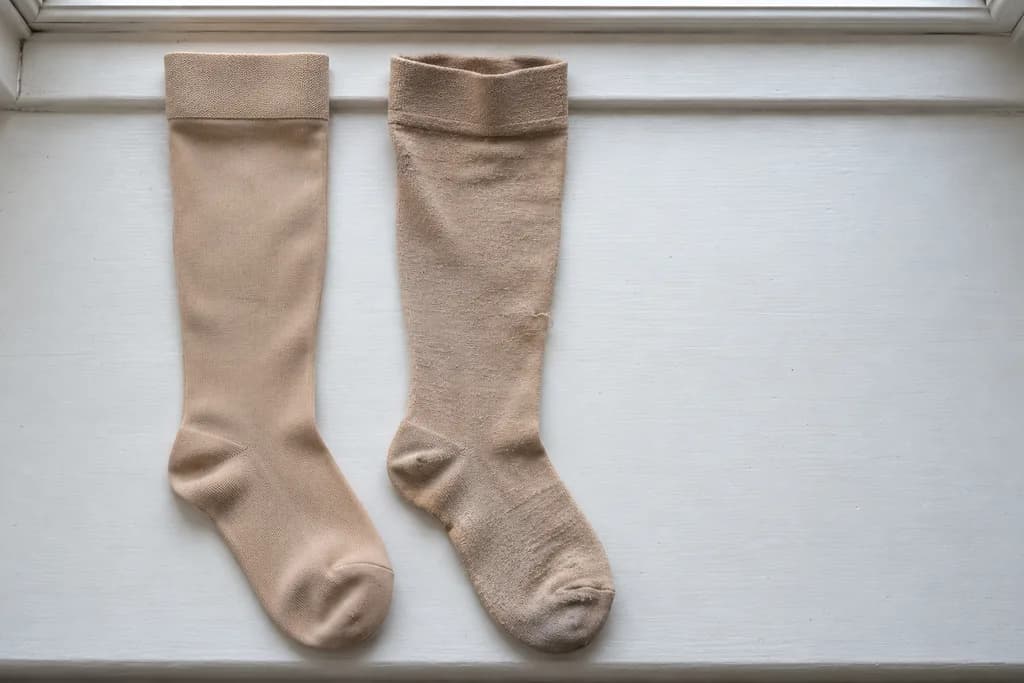 Zwei beige Kompressionsstrümpfe nebeneinander auf einer weißen Fensterbank, links ein neuer mit glattem Gestrick, rechts ein getragener mit Knötchen, grauem Fußteil und schlaffem Bund.