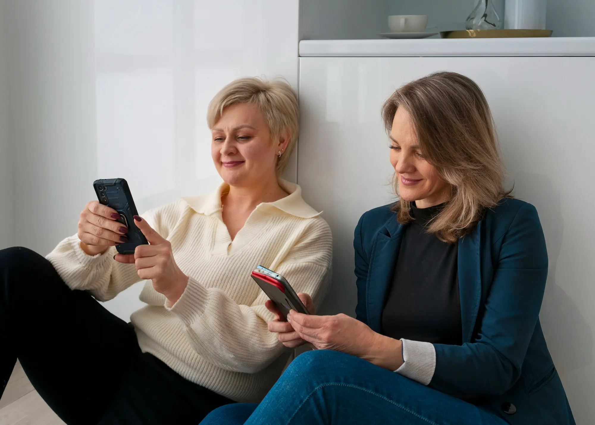 Zwei Frauen Ende 50 sitzen entspannt auf dem Boden an einer weißen Wand gelehnt und schauen jeweils konzentriert auf ihre Smartphones. Eine trägt einen cremefarbenen Pullover, die andere einen petrolblauen Blazer. Helles, modernes Zuhause, freundliche Stimmung.