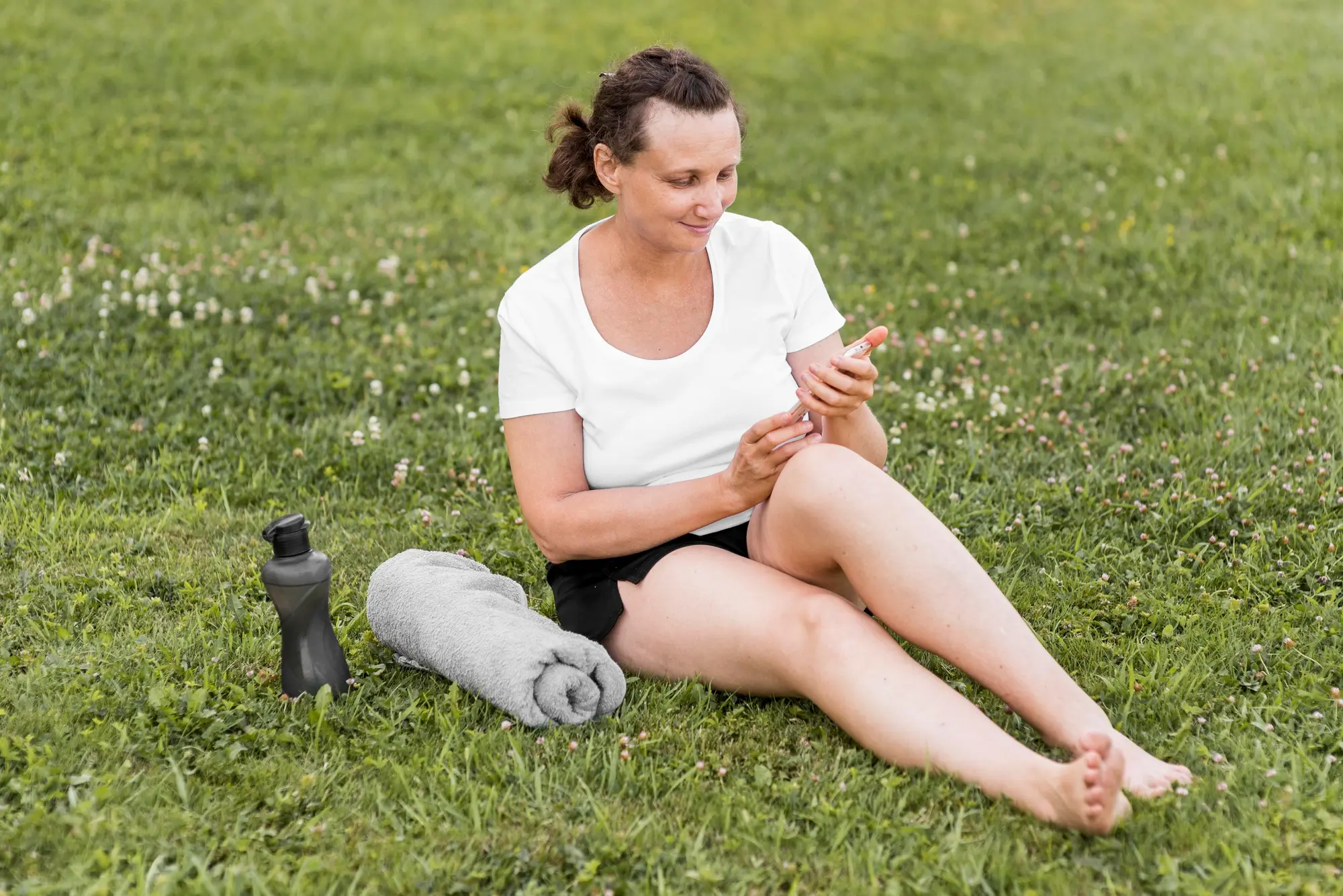 Frau Ende 50 mit dunklem Pferdeschwanz sitzt entspannt im weißen T-Shirt und schwarzen Sportshorts auf einer Wiese im Park, schaut auf ihr Smartphone. Neben ihr eine schwarze Trinkflasche und ein zusammengerolltes graues Handtuch, weiches Tageslicht und niedriger Blickwinkel.