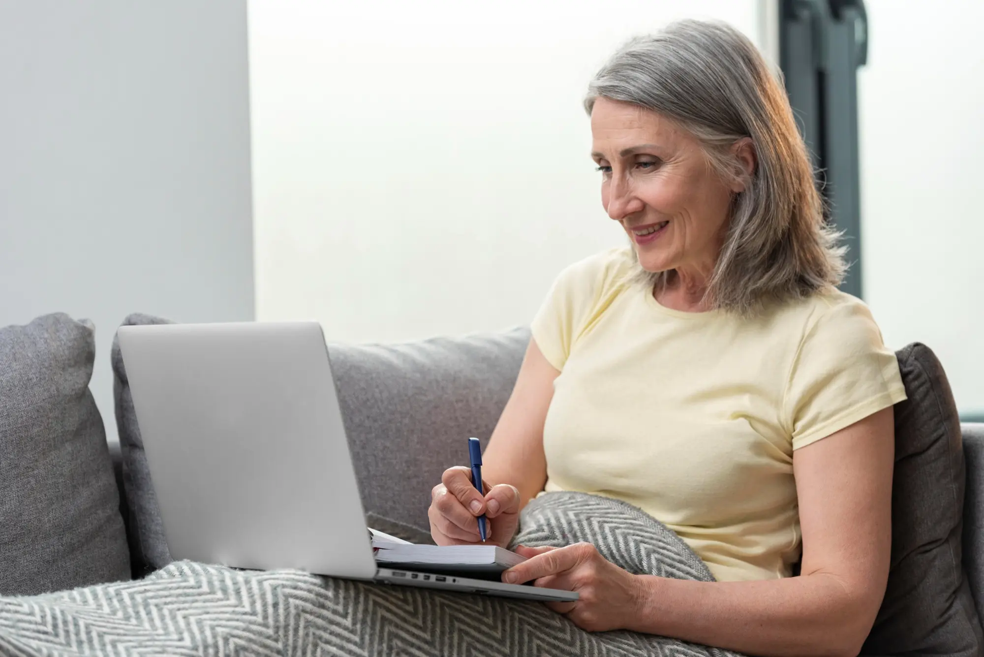 Lächelnde Frau Ende 50 mit silbergrauem Haar sitzt auf einem grauen Sofa, hat einen Laptop auf dem Schoß und macht sich konzentriert Notizen in einem aufgeschlagenen Heft. Helles, freundliches Wohnzimmer mit weichem Tageslicht.