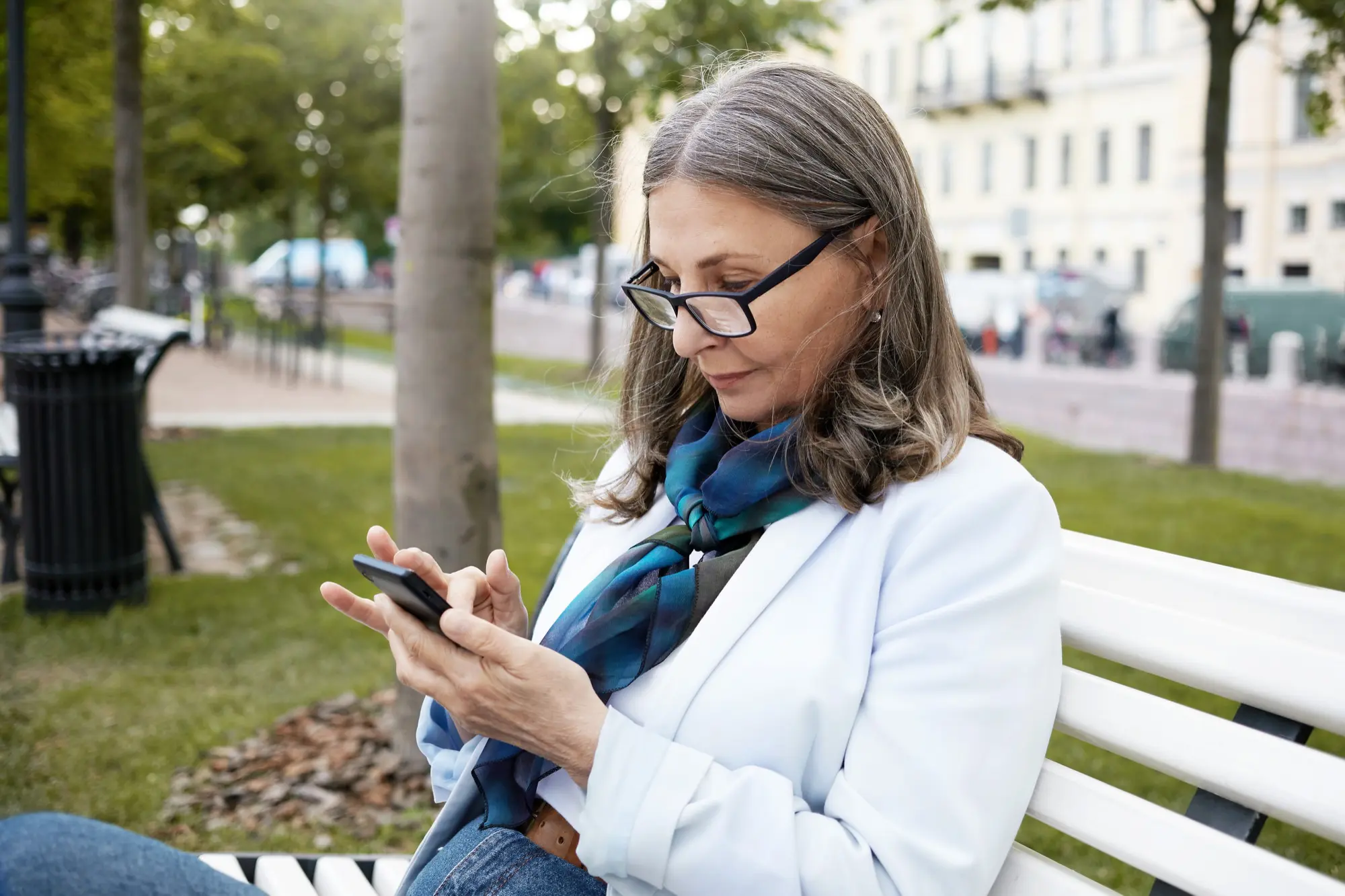 Frau Ende 50 mit silbergrauem Haar und Brille sitzt auf einer weißen Parkbank in der Stadt und schaut konzentriert auf ihr Smartphone. Helle Blazerjacke, blau-grüner Schal, im Hintergrund Bäume, Bürgersteig und Häuserfassaden.