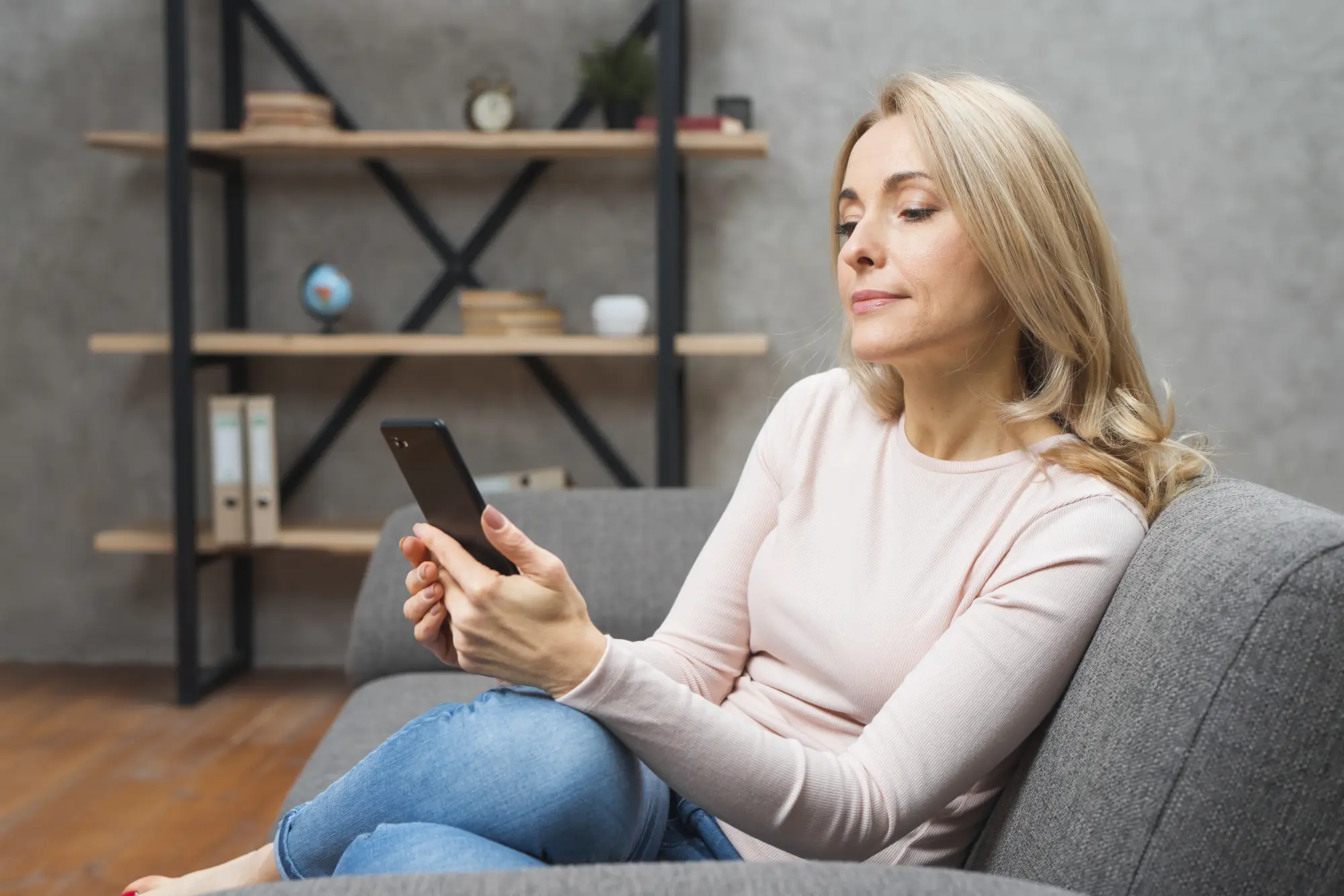 Frau Mitte 40 mit langen blonden Haaren sitzt entspannt auf einem grauen Sofa und schaut auf ihr Smartphone. Sie trägt einen rosafarbenen Pullover und eine Jeans, im Hintergrund ein offenes Regal mit Büchern und Deko.
