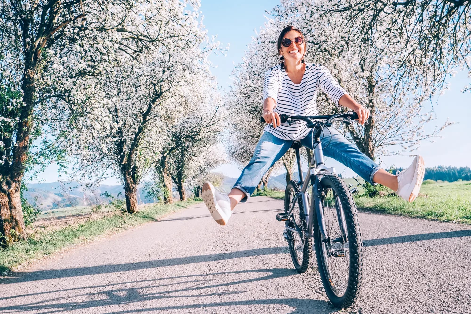 A smiling middle-aged woman cycling along a sunny country road lined with white-blossoming trees. She wears a striped shirt, jeans and sunglasses, and kicks both legs out to the sides with joy. Bright spring light, carefree mood.