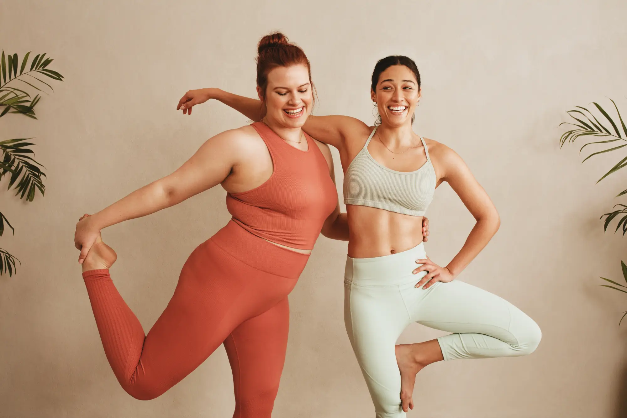 Zwei lachende Frauen in Sportkleidung stehen vor einer warmen beigen Wand und machen gemeinsam Yoga-Übungen. Eine steht in der Baumhaltung, die andere balanciert auf einem Bein. Grüne Zimmerpflanzen seitlich, entspannte und stärkende Atmosphäre.