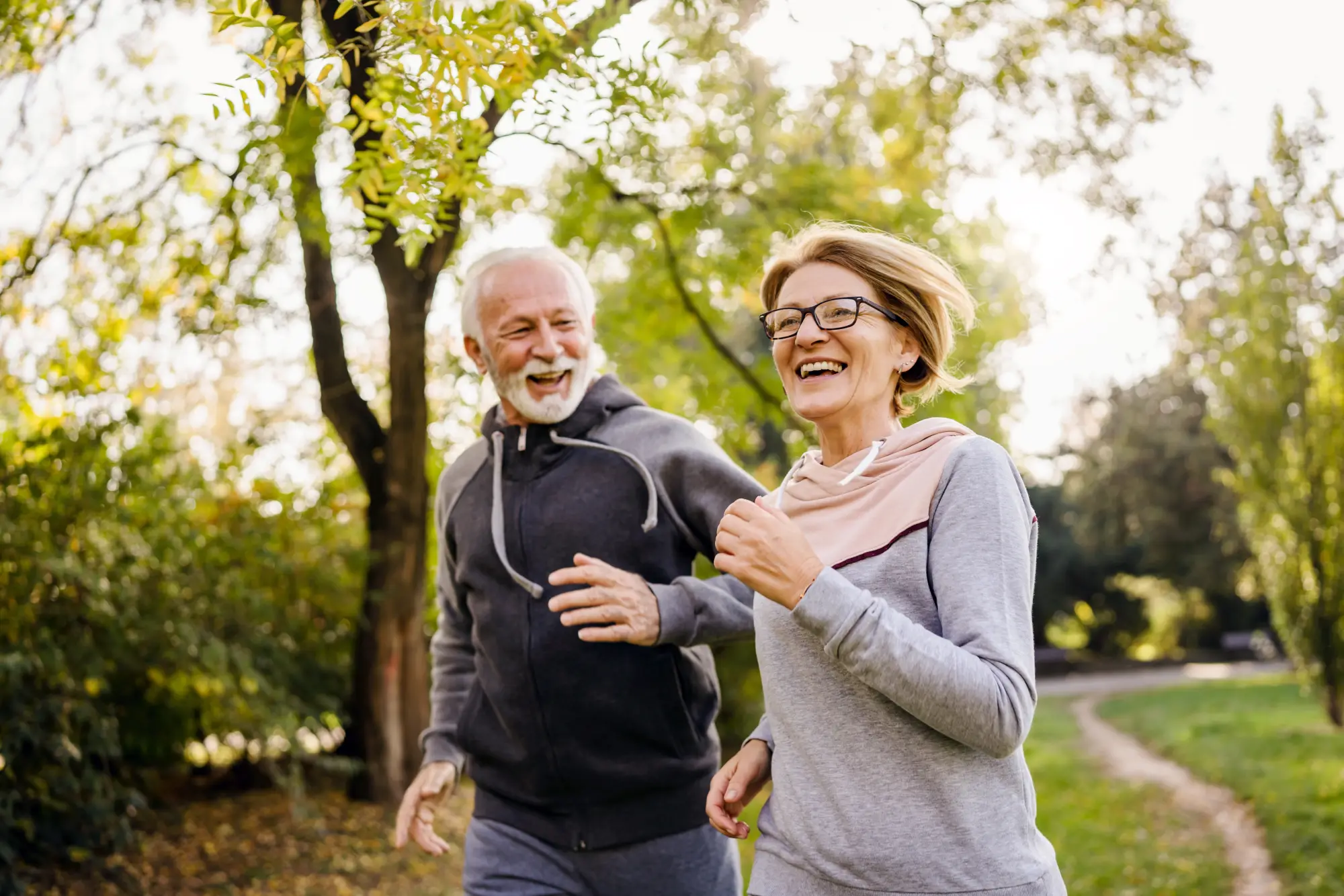 Ein lachendes Paar Anfang 60 joggt gemeinsam an einem sonnigen Herbsttag durch einen Park. Beide tragen sportliche Kapuzenpullover, im Hintergrund grüne Bäume und ein geschwungener Weg. Lebensfrohe, aktive Stimmung.