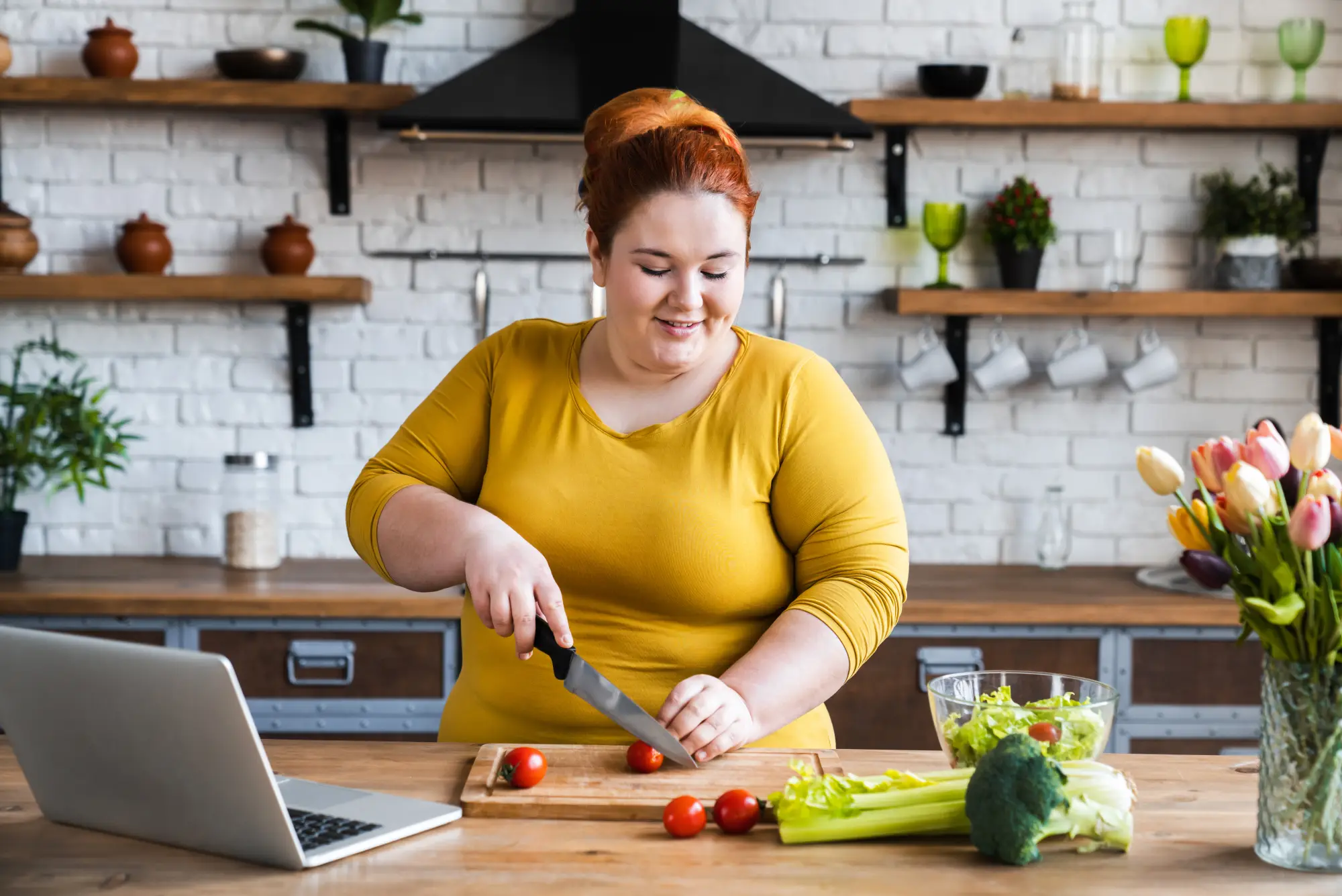 Eine lächelnde Frau mit rotem Haar schneidet in einer hellen Küche frische Tomaten auf einem Holzbrett. Vor ihr ein aufgeklappter Laptop, daneben Salat, Sellerie und Brokkoli sowie ein Strauß Tulpen. Entspannte, gesunde Alltagsstimmung.