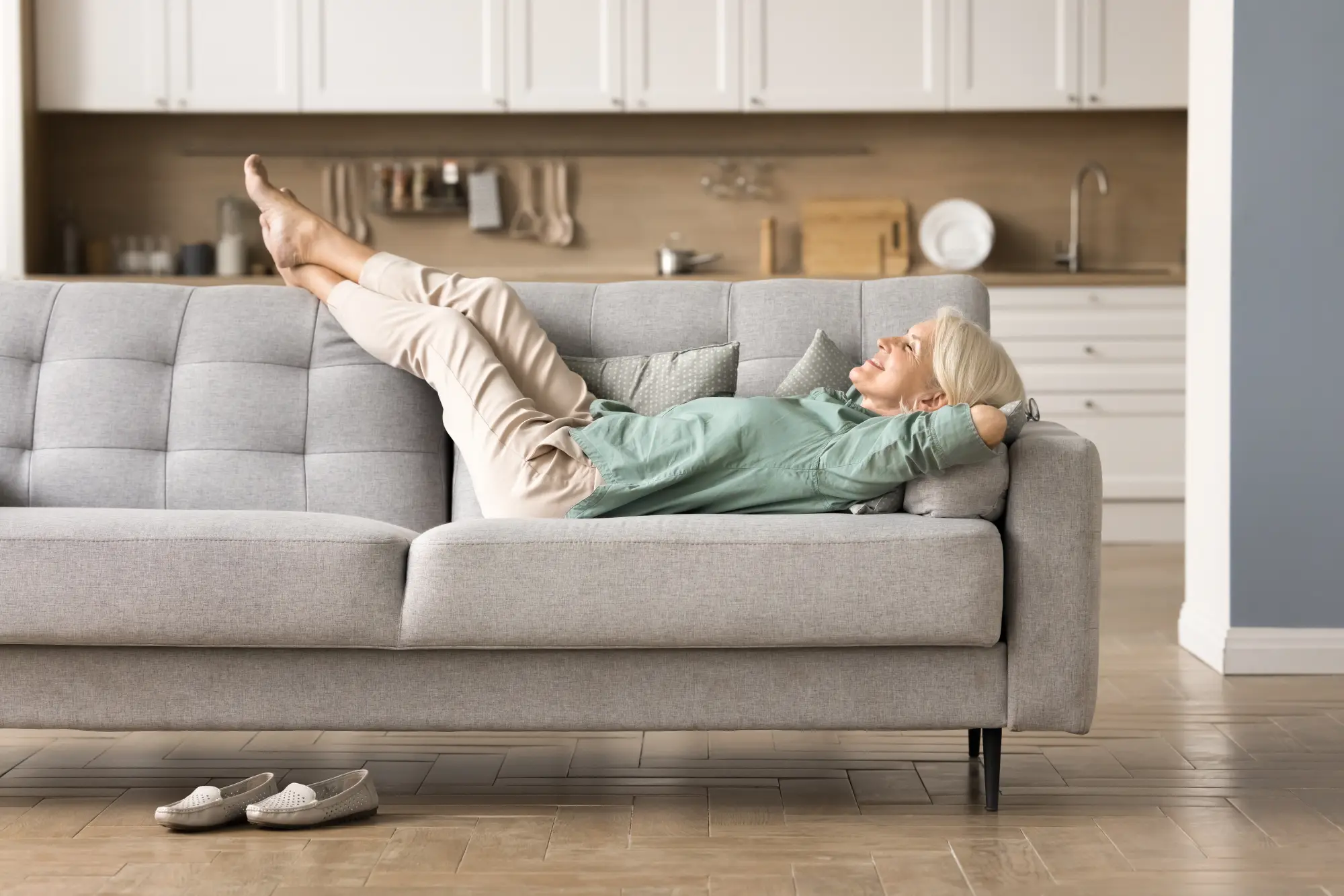Frau Anfang 60 mit silbergrauem Haar liegt entspannt auf einem grauen Sofa, die Beine hochgelegt und die Hände hinter dem Kopf verschränkt. Grüne Bluse, cremefarbene Hose, barfuß, Slipper stehen vor dem Sofa. Im Hintergrund eine helle Wohnküche, ruhige Nachmittagsstimmung.