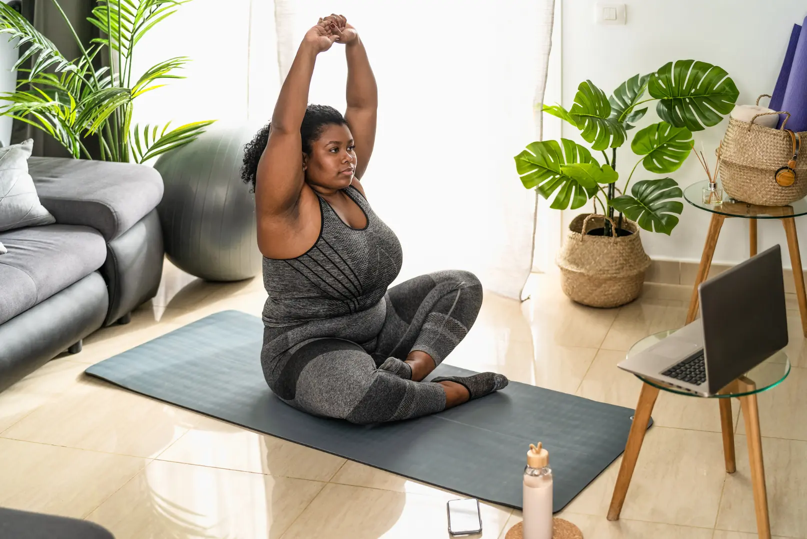 Frau auf einer Yogamatte in einem hellen Wohnzimmer streckt beide Arme über den Kopf, neben ihr ein Laptop mit einer angeleiteten Übung. Zimmerpflanzen, ein Gymnastikball und eine Wasserflasche im Raum, ruhige Trainingsstimmung zu Hause.