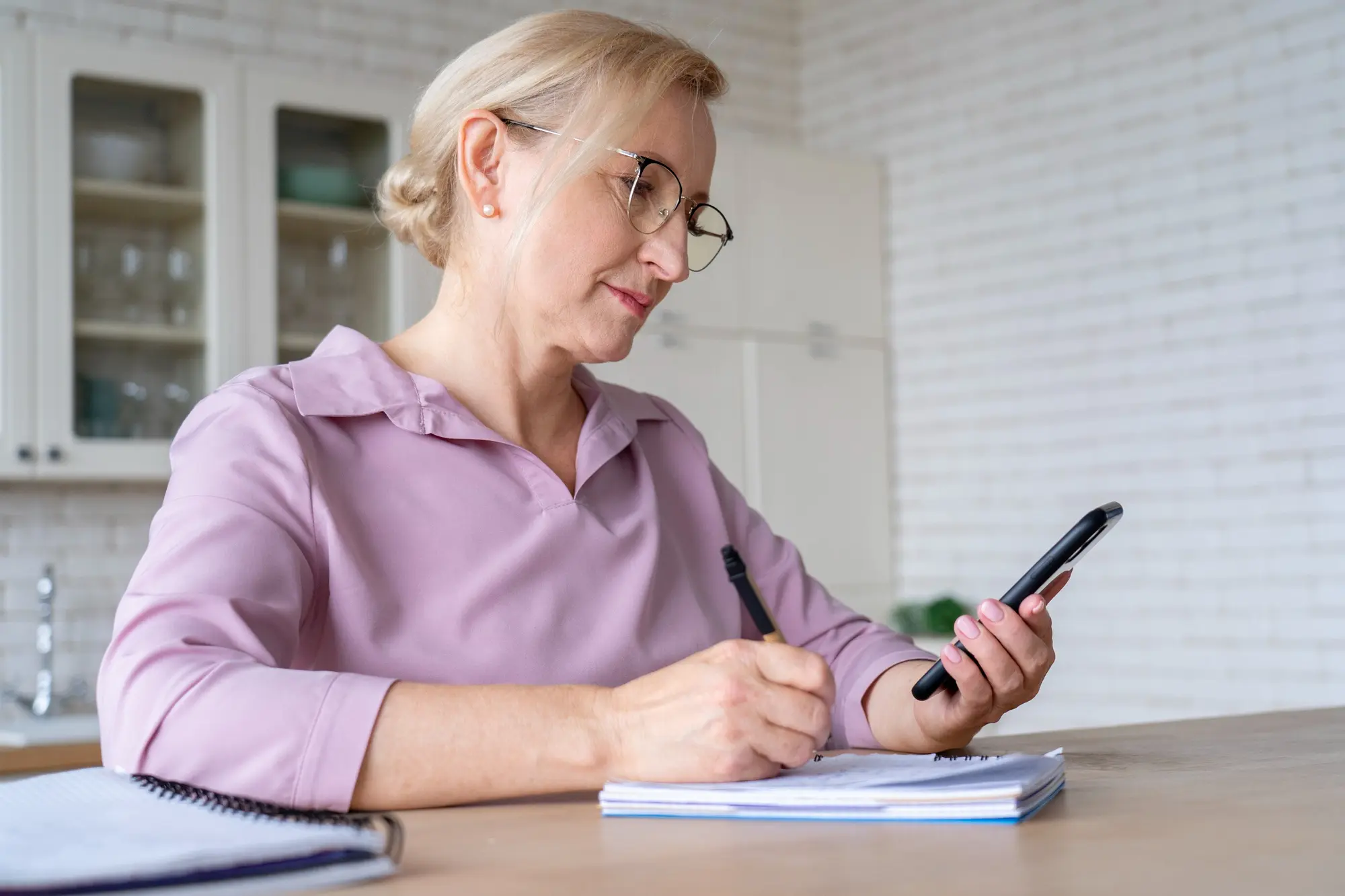 Frau Ende 50 mit blondem Haar und Brille in einer rosafarbenen Bluse sitzt in einer hellen Küche an einem Tresen, hält ihr Smartphone in der Hand und notiert etwas in einem Spiralblock. Weitere Notizbücher auf der Arbeitsfläche, weiches Tageslicht.