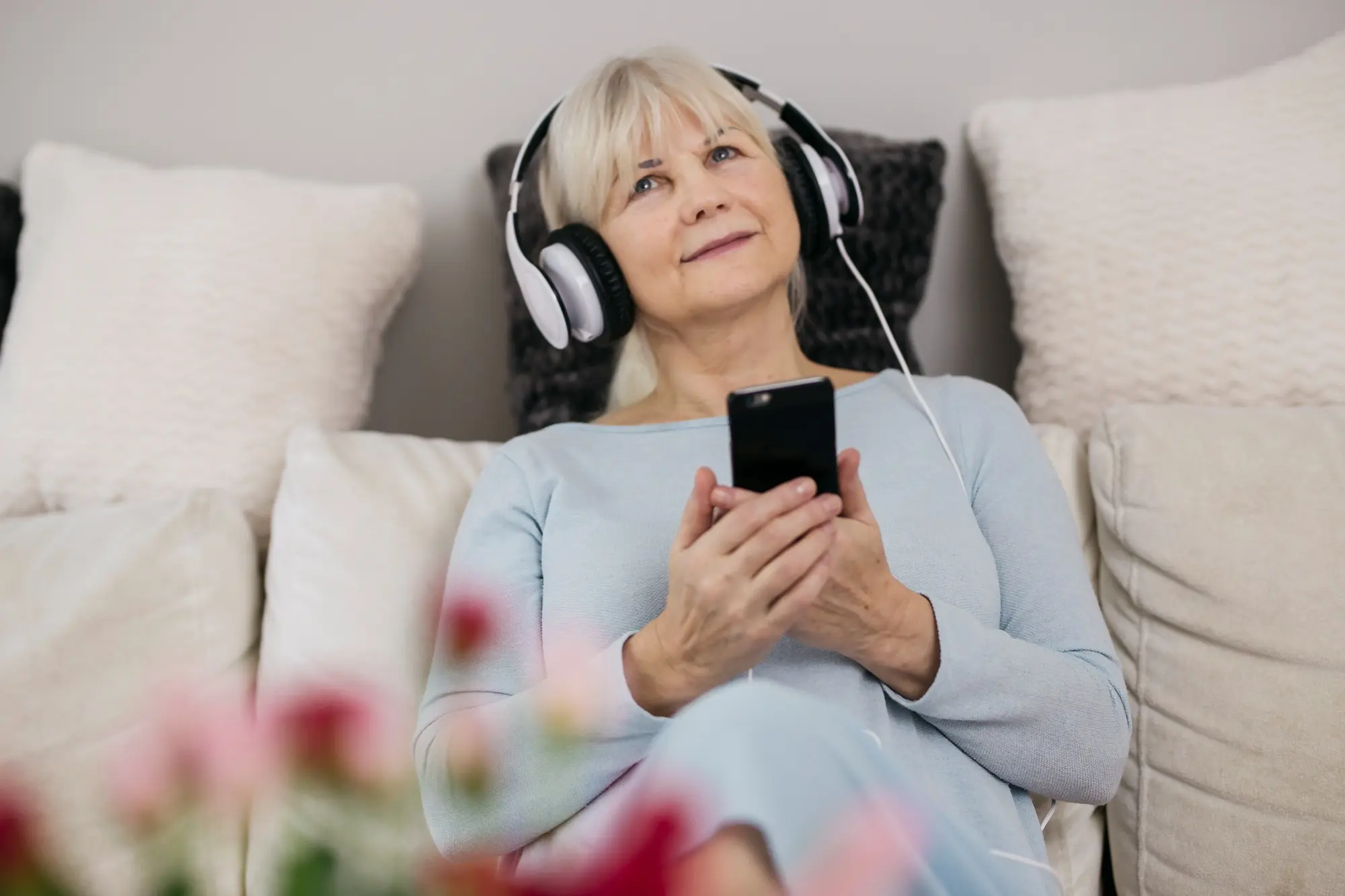 Frau Anfang 60 mit silbergrauem Haar in hellblauem Oberteil sitzt entspannt auf einem Sofa, trägt Over-Ear-Kopfhörer und hört auf ihrem Smartphone etwas. Helle Kissen, im Vordergrund unscharfe rosa Blüten, ruhige Stimmung.